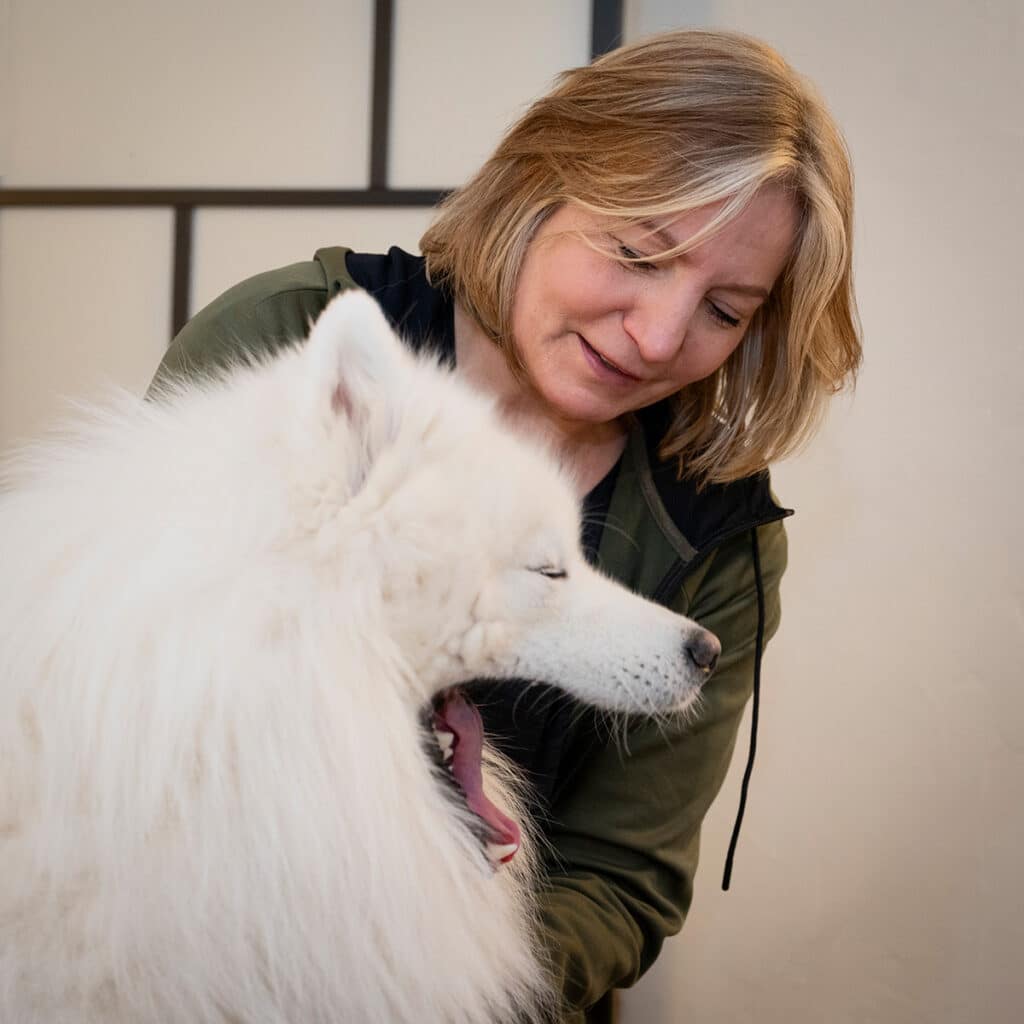 Suzanne tittar kärleksfullt på en fridfull vit, fluffig hund, som verkar gäspa eller rösta och delar en stund av sällskap och förnöjsamhet för hundar.
