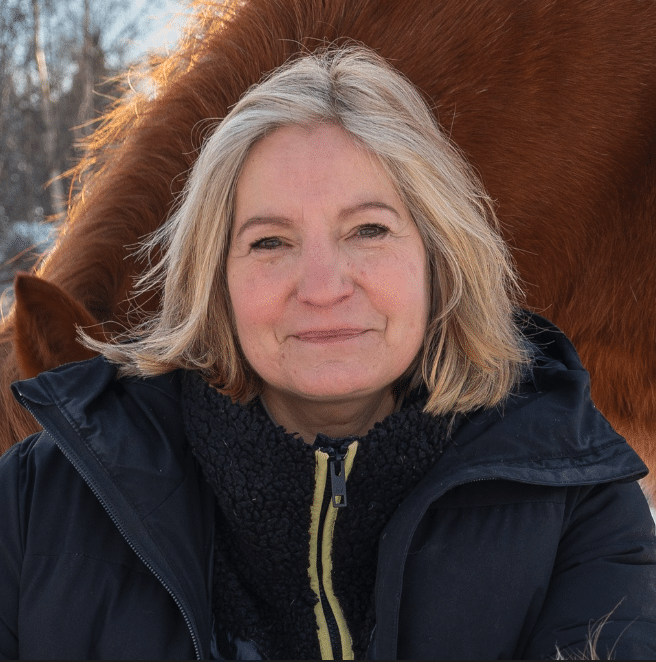 En leende Suzanne med blont hår, klädd i en vinterjacka, står utomhus med en mjuk bakgrund som antyder närvaron av en häst för brainwork.